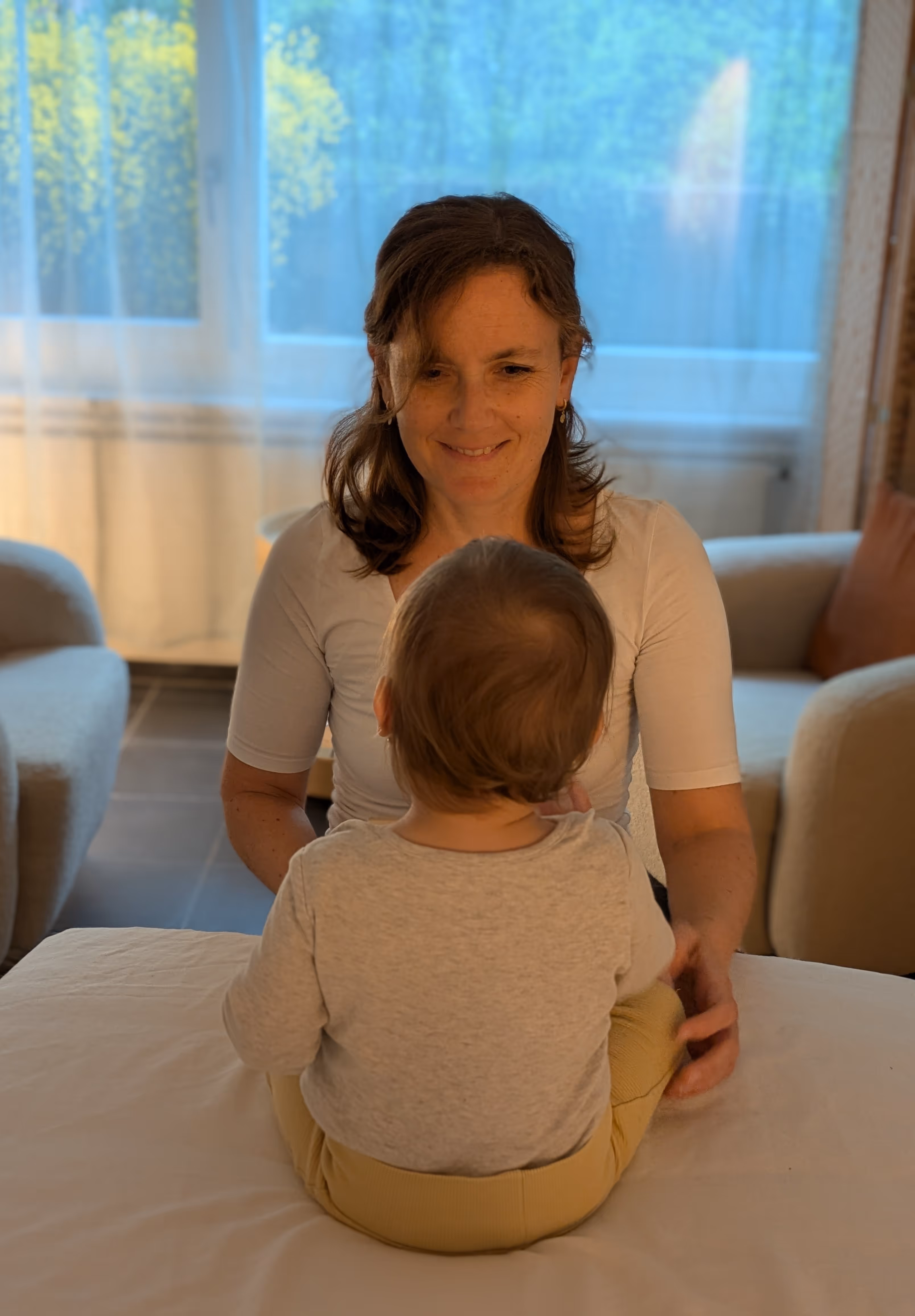 Frau legt Baby in weißen Kleid auf blauem Tuch auf eine Liege in einem hellen Raum.