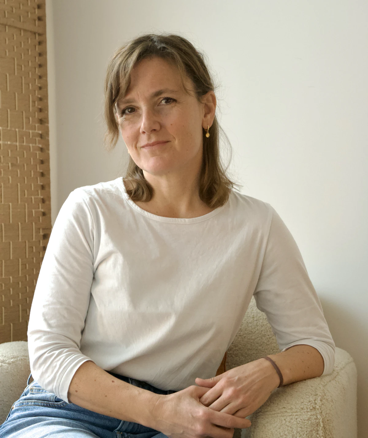 Frau sitzt entspannt in einem hellen Raum auf einem hellen weichen Sessel, trägt ein weisses Langarmshirt und blaue Jeans.