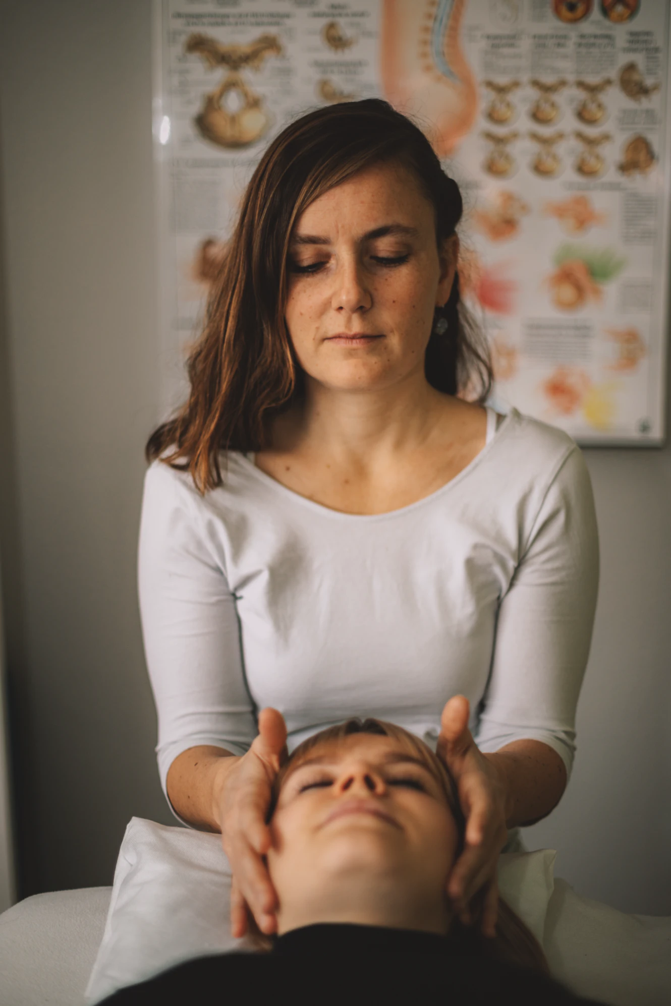 Frau in weissem Oberteil berührt den Kopf einer liegenden Person in einem Behandlungsraum mit anatomischem Poster im Hintergrund.